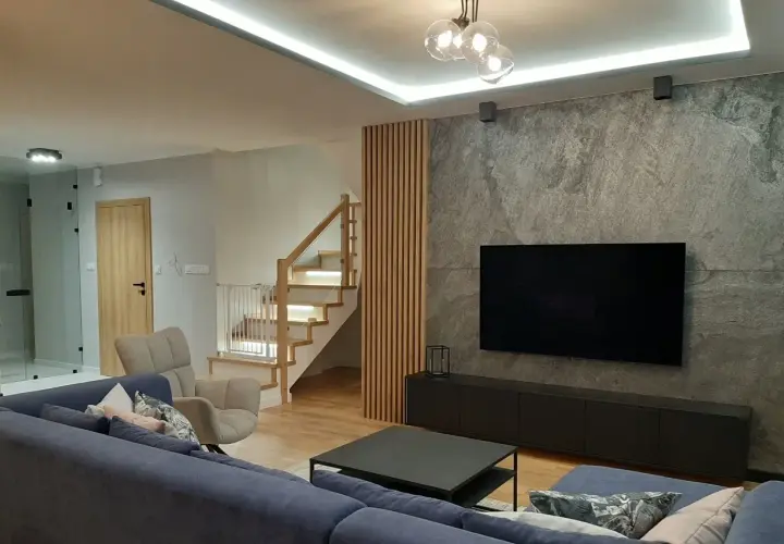 Nowoczesny salon z szarą ścianą i telewizorem. Drewniane lamele idealnie komponują się ze schodami i podłogą, tworząc ciepłą atmosferę.