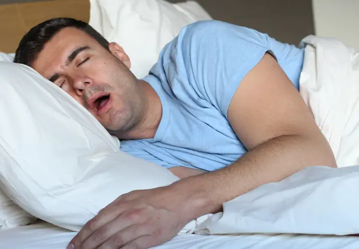 Uomo dorme a bocca aperta, spiegando perchè si sbava quando si dorme. La sua testa è appoggiata su un cuscino bianco.