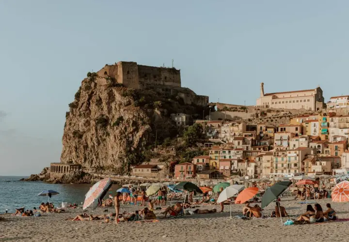 A sunny beach with people relaxing under umbrellas, overlooking a cliffside town with a castle. This view is a perfect destination for those wondering how to get to Calabria, Italy.