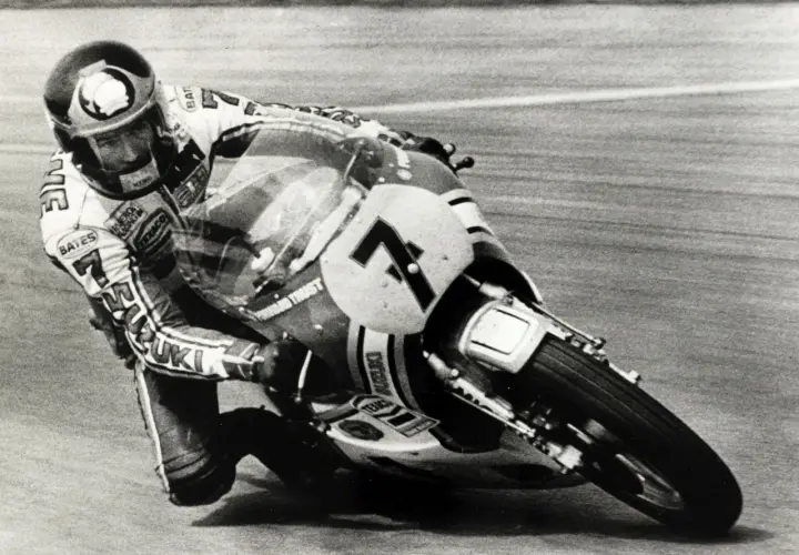 Wyścigowy motocykl Suzuki z numerem 7 pochylony w zakręcie, z zawodnikiem w kasku. To fragment historii motocykla.