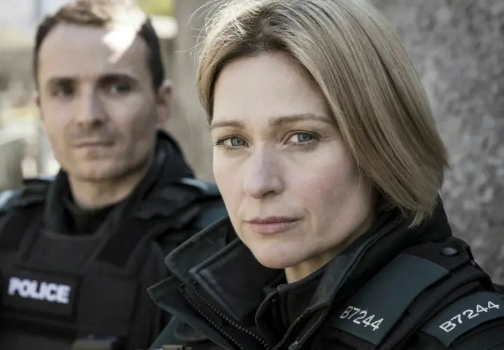 Två poliser i uniform, en kvinna i förgrunden med intensiv blick, bakgrunden svagt belyst av blåa ljus.