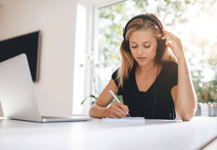 Mujer con auriculares escribe en un cuaderno, con luz natural entrando por la ventana. La luz suave es ideal, preguntándose cuál es la mejor luz para estudiar.