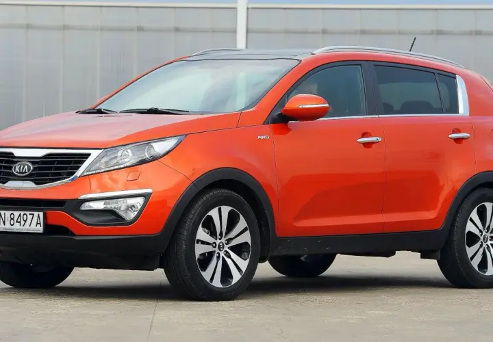 Pomarańczowy Kia Sportage 2015 z napędem AWD, zaparkowany na betonowym placu przed budynkiem z przeszkloną fasadą.