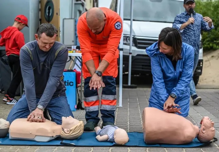 Ratownik medyczny w pomarańczowym stroju nadzoruje ćwiczenia resuscytacji na fantomach. Uczestnicy uczą się ratować życie.