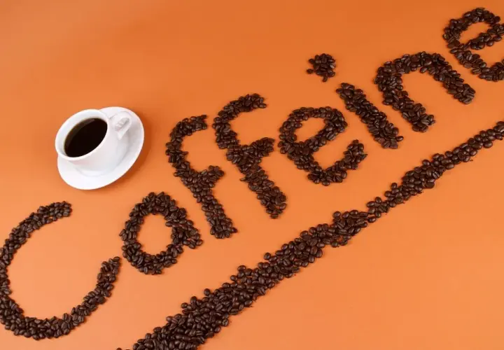 Ziarna kawy ułożone w napis "caffeine" obok filiżanki kawy. Zastanawiasz się, ile kofeiny ma łyżeczka kawy mielonej?