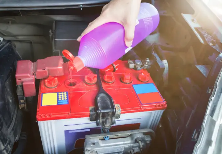 Ręka wlewa fioletowy płyn, prawdopodobnie elektrolit do akumulatora, do czerwonej obudowy akumulatora samochodowego.