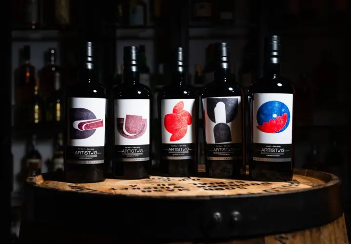 Cinq bouteilles de blend whisky Artist-13, ornées d'étiquettes artistiques colorées, reposent sur un fût de whisky.