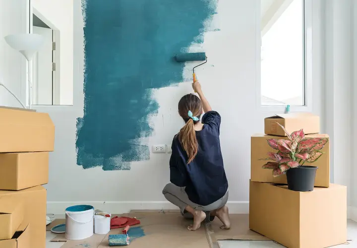 Mujer pintando una pared de azul, rodeada de cajas. Es la mejor época para pintar la casa y darle un nuevo aire.