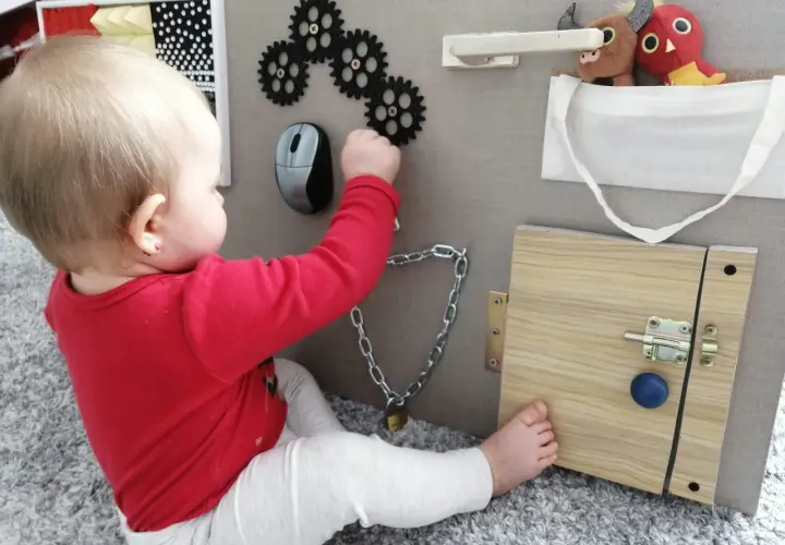 Dziecko bawi się przy tablicy manipulacyjnej diy: zębatki, myszka, zamek, kłódka i pluszaki.