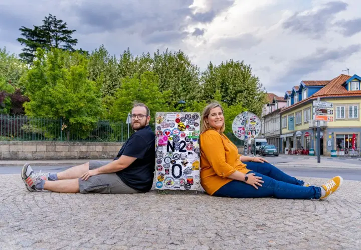 Pareja sentada de espaldas en un poste con pegatinas, celebrando su viaje por la Estrada Nacional 2 en Portugal.