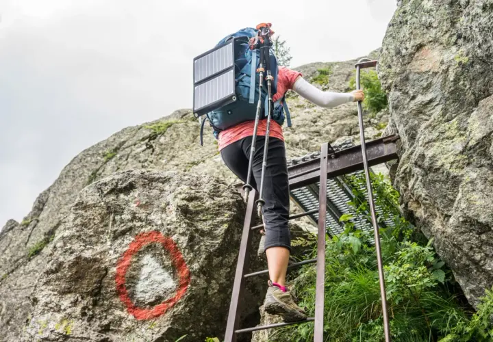 A hiker ascends metal ladders on the Tour du Mont Blanc, using trekking poles and a solar panel on their backpack.