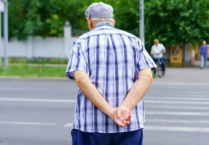 Starszy pan w kratowanej koszuli i czapce, trzymając ręce za plecami, czeka na zielone światło na pasach.