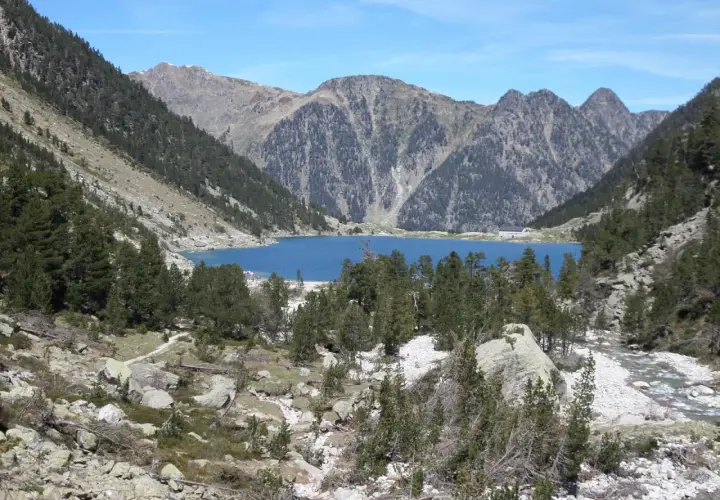 Lago azul rodeado de montañas y pinos, con un pequeño refugio en la orilla. El refugio Oulettes de Gaube es un destino popular para excursionistas.