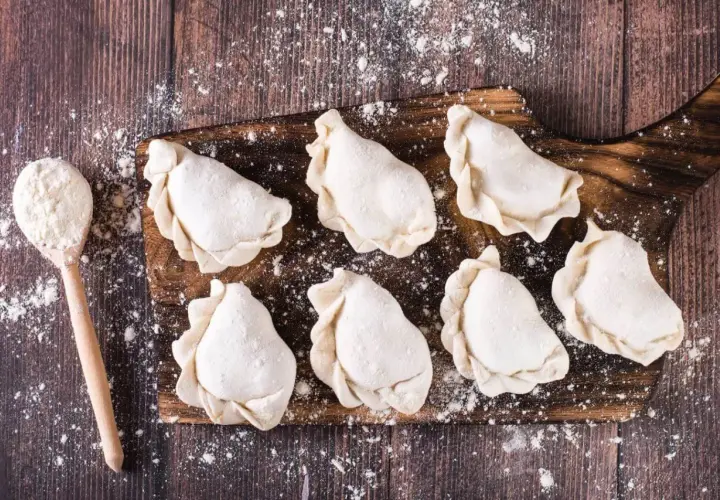 Surowe pierogi na desce, gotowe do przechowywania w lodówce. Obok łyżka z mąką.