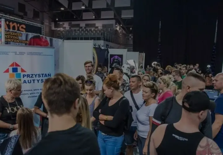 Ludzie na konwencie tatuażu, gdzie widoczny jest symbol autyzmu na banerze "Przystań Autys".