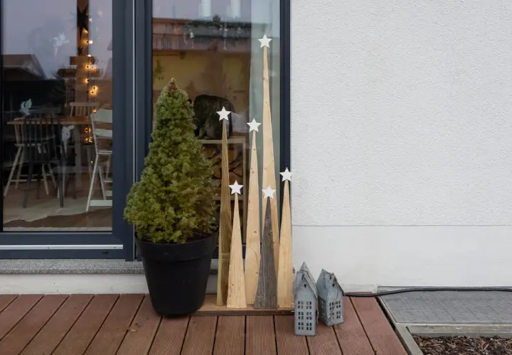 Selbstgemachte Weihnachtsdeko außen: Tannenbaum im Topf, Holz-Tannenbäume mit Sternen und kleine Lichterhäuser auf einer Holzterrasse.