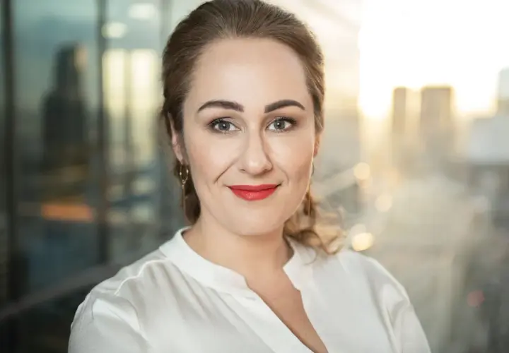 Portretowa fotografia bizneswoman z uśmiechem, w białej bluzce, na tle rozmazanego miasta.