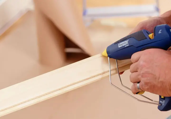 Här limmas golvlister med en limpistol. En person applicerar smältlim på en trälist för att fästa den.