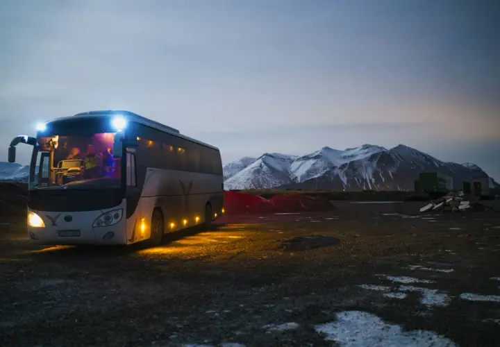 Autobus z zapalonymi światłami na tle ośnieżonych gór o zmierzchu.