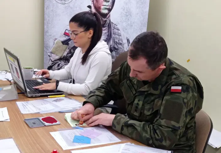 Żołnierz w mundurze pieczętuje dokumenty, podczas gdy kobieta pracuje przy laptopie. To część procesu kwalifikacji wojskowej.