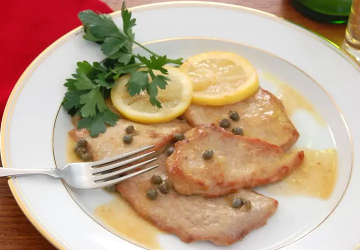 Piccata de veau, tendre escalope de veau sauce citronnée, câpres et persil frais, sur une assiette blanche bordée d'or.
