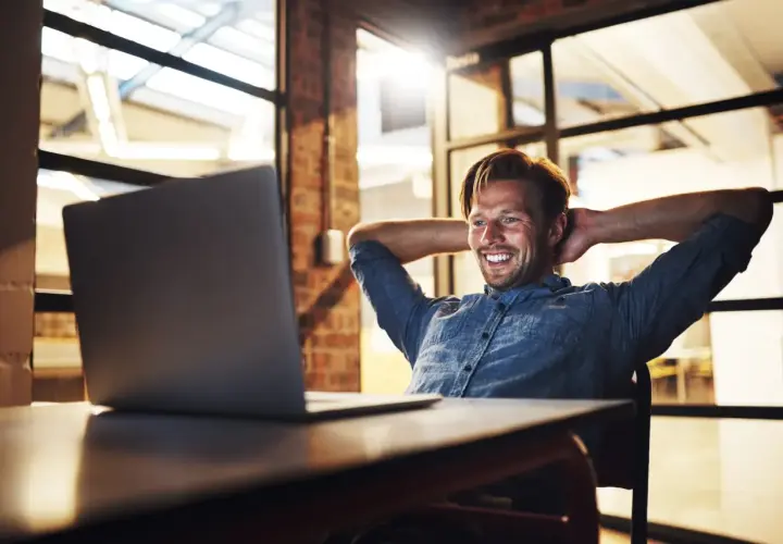 Uśmiechnięty mężczyzna w back office, z rękami za głową, patrzy na laptopa.