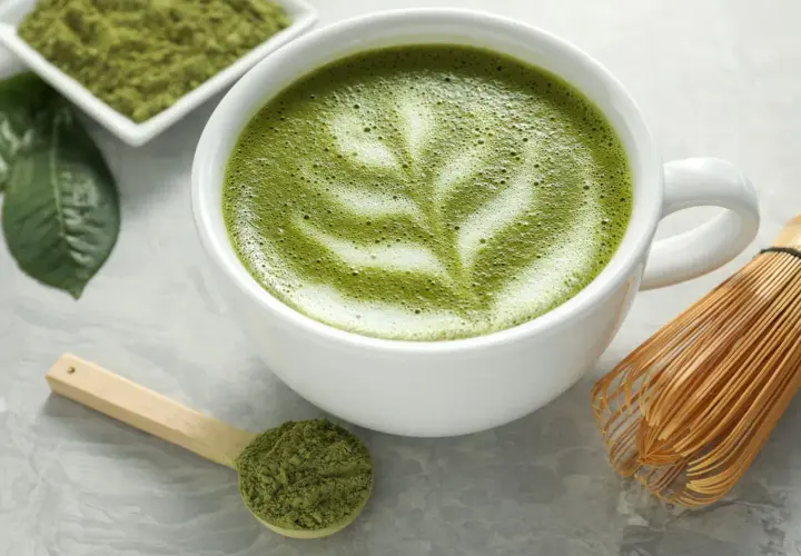 Zielona matcha w filiżance z pianką, obok miarka z proszkiem i bambusowa miotełka. Czy matcha ma kofeinę? Tak, zawiera ją!