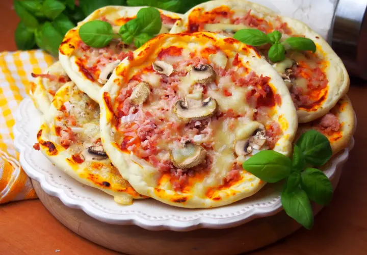 Szybkie mini pizze z szynką i pieczarkami, ozdobione świeżą bazylią, kuszą apetycznym wyglądem.
