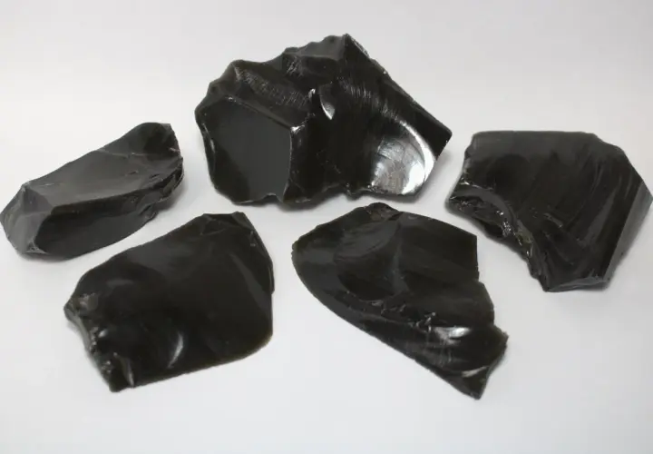 Fünf scharfkantige Stücke glänzenden, schwarzen Obsidiansteins liegen auf weißem Untergrund.