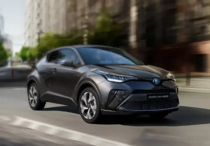 Ciemnoszary Toyota C-HR Hybrid w ruchu miejskim, z rozmytym tłem budynków i drzew.