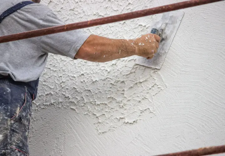 Un operaio applica intonaco rustico con una spatola, creando una texture grezza e irregolare su una parete esterna.