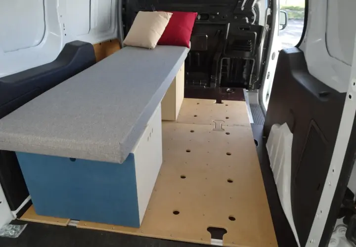 Aménagement intérieur d'un petit fourgon : banquette grise avec coussins, caisson de rangement bleu et blanc, plancher en bois. Prêt pour l'aventure !