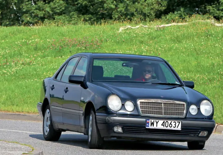 Czarny Mercedes W210 z silnikiem om606 jedzie po drodze, za nim zielone, kwieciste wzgórze.