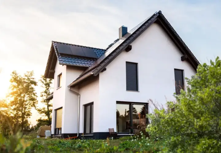 Nowy biały dom z panelami słonecznymi na dachu. Słońce zachodzi za drzewami. Idealny czas na wycenę budowy domu.
