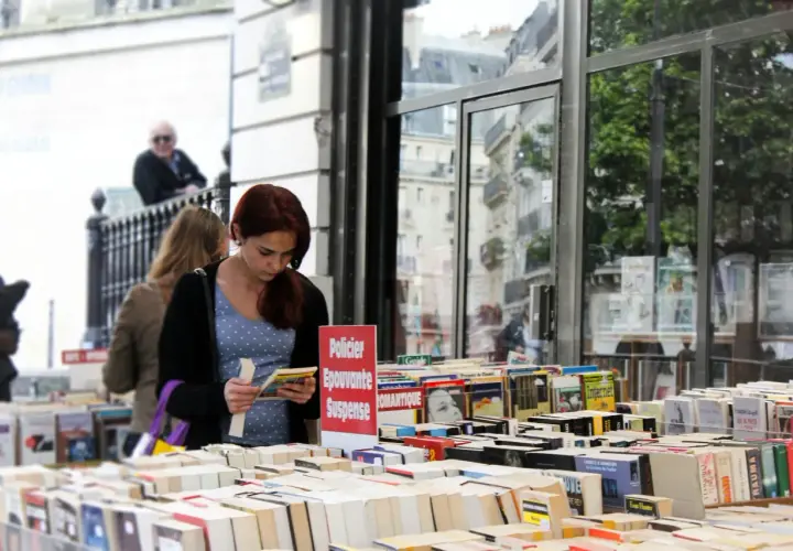 Une jeune femme choisit un livre dans une librairie en plein air, devant une vitrine reflétant des bâtiments parisiens. Une sélection de livres, dont des romans policiers et de suspense, est exposée, rappelant une maison d'édition jeunesse.
