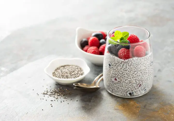 Pudding chia z malinami i borówkami, ozdobiony miętą, obok miseczki z nasionami chia.