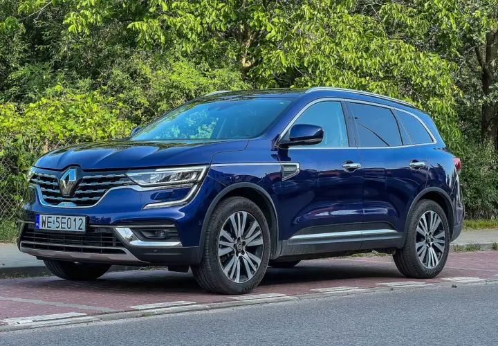 Granatowy Renault Koleos stoi na czerwonej kostce brukowej, otoczony zielenią.
