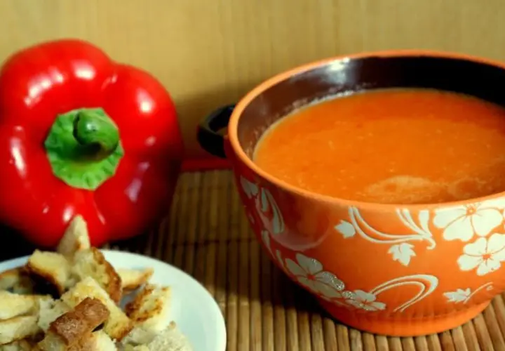 Aksamitny zupa krem z papryki, podana z chrupiącymi grzankami. Obok leży świeża, czerwona papryka.