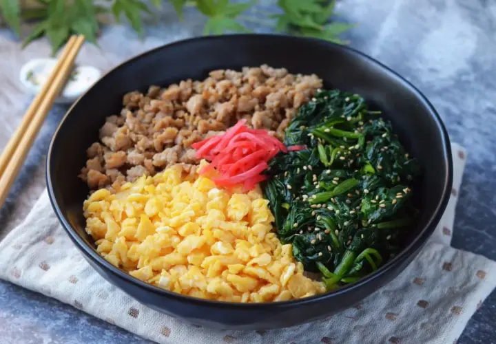 A colorful sanshoku don bowl with scrambled eggs, seasoned ground meat, and spinach, topped with pickled ginger.