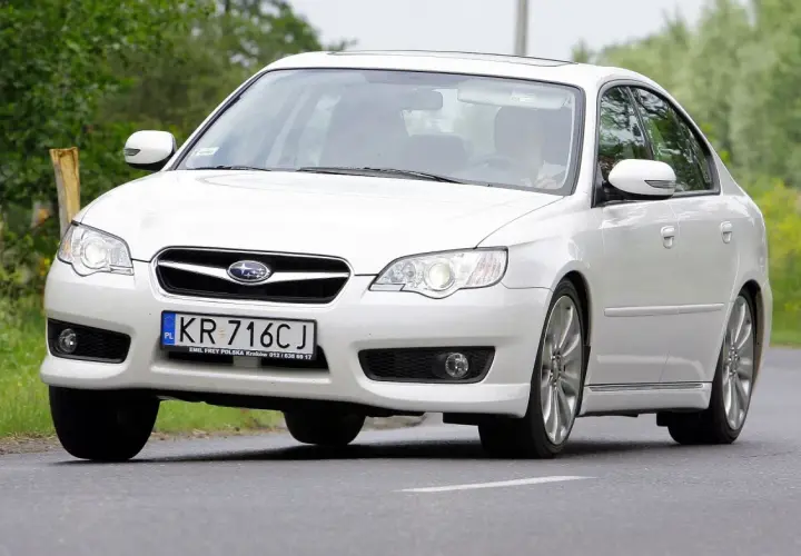 Białe Subaru Legacy z polskimi tablicami KR 716CJ jedzie po drodze wśród zieleni.