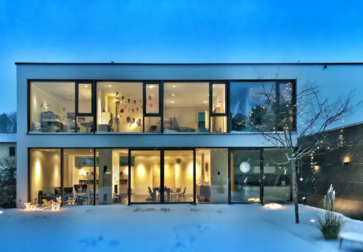 Casa moderna con grandes ventanales iluminados, mostrando interiores acogedores. La nieve cubre el exterior, y luces navideñas decoran un árbol. ¡2500 lumens cuanto ilumina!