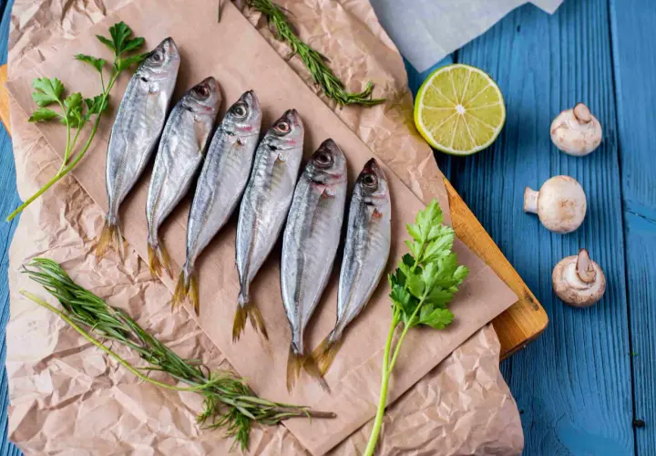 Seis jureles pescado frescos, listos para cocinar, acompañados de perejil, romero, limón y champiñones sobre papel de estraza y tabla de madera.