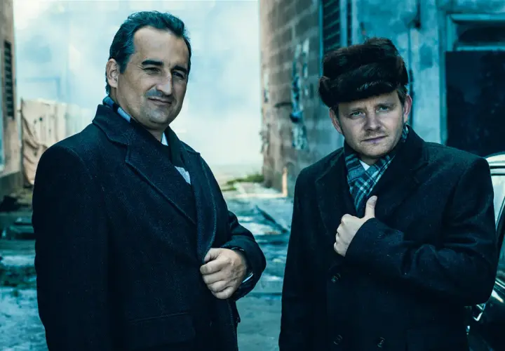 Obsada filmu "Gierek": dwaj mężczyźni w płaszczach i szalikach, jeden w futrzanej czapce, stoją obok czarnego samochodu.