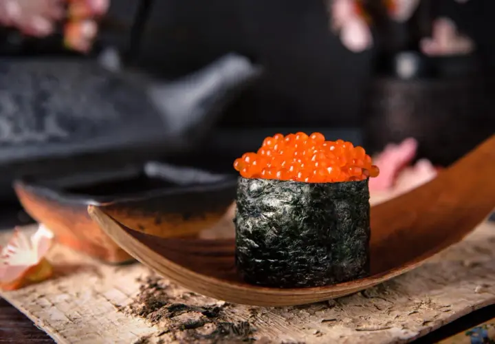 Wykwintny gunkan sushi z łososiem ikra, owinięty w ciemne nori, prezentowany na drewnianej łódce.