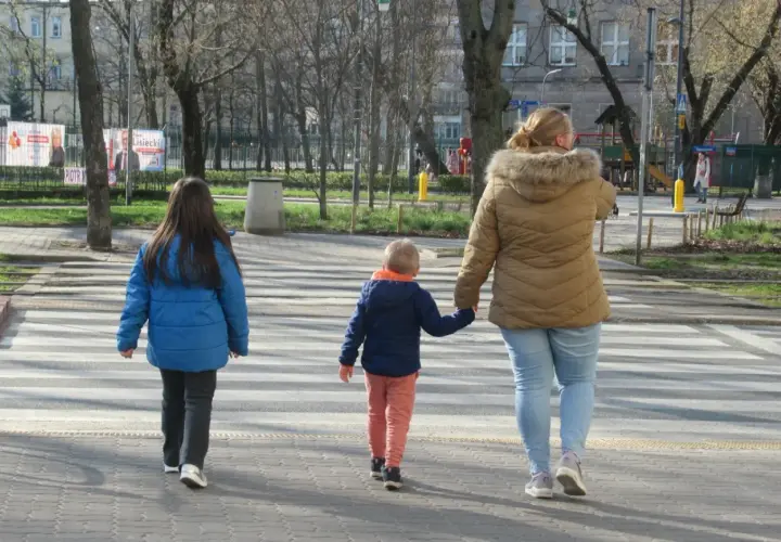 Rodzina przechodzi przez pasy. Kobieta trzyma za rękę małego chłopca, a obok idzie starsza dziewczynka. Szczególna ostrożność kiedy przechodzimy przez jezdnię.