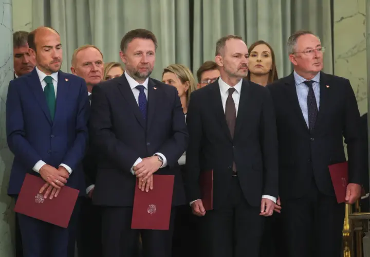 Mężczyźni w garniturach, być może posłowie, stoją w rzędzie, trzymając czerwone dokumenty.