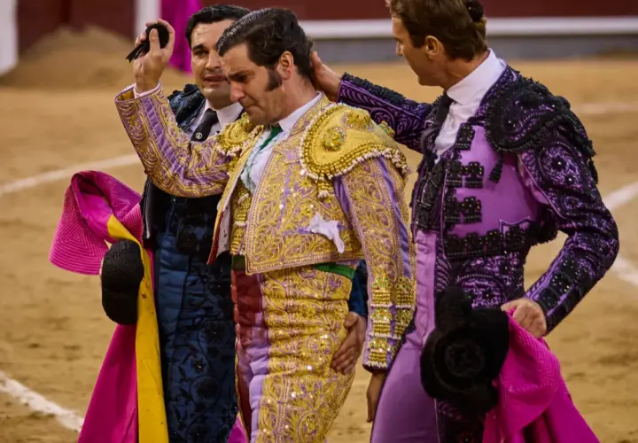 Tres toreros con trajes de luces, uno con coleta de torero, son felicitados en la arena.