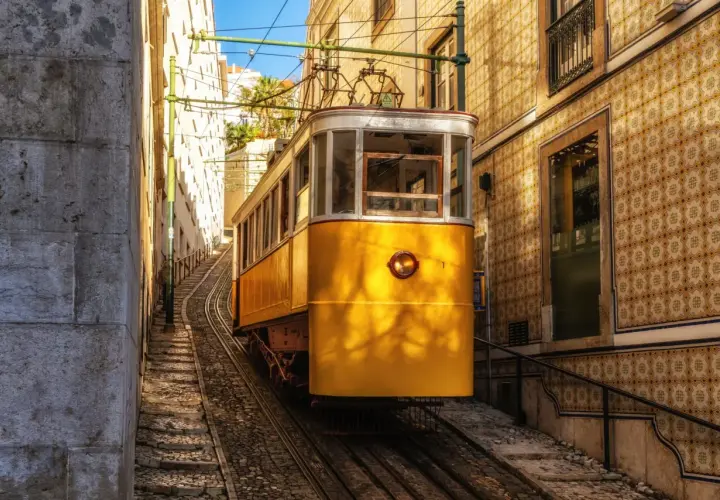 Żółty tramwaj wspina się po stromych torach w wąskiej uliczce Lizbony. W tle widać kamienne budynki pokryte tradycyjnymi płytkami azulejo.
