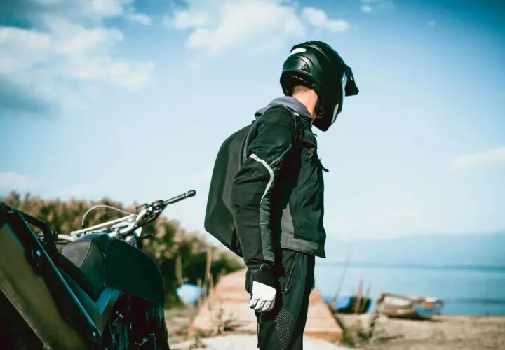 Motociclista con casco negro, chaqueta y guantes blancos, junto a su moto. El tejido de poliéster 600d vs Cordura es ideal para su aventura.