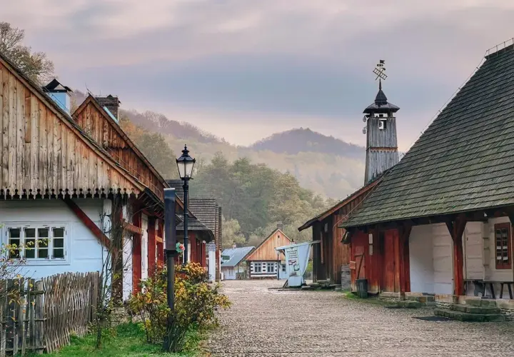 Sanok skansen: urokliwa, brukowana uliczka z drewnianymi domami i wieżą z wiatrowskazem, otoczona mglistymi wzgórzami.